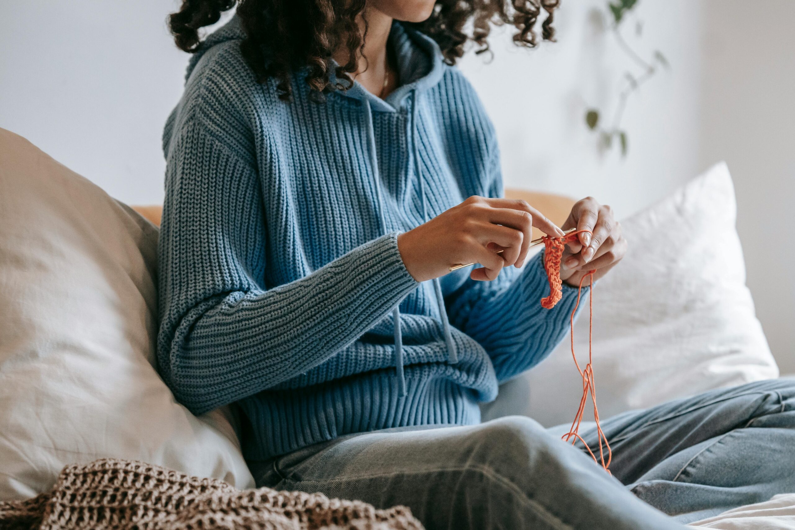 Crop anonymous female in knitted blue hoodie and jeans sitting on sofa surrounded with pillows and crocheting with orange thread and crochet hook